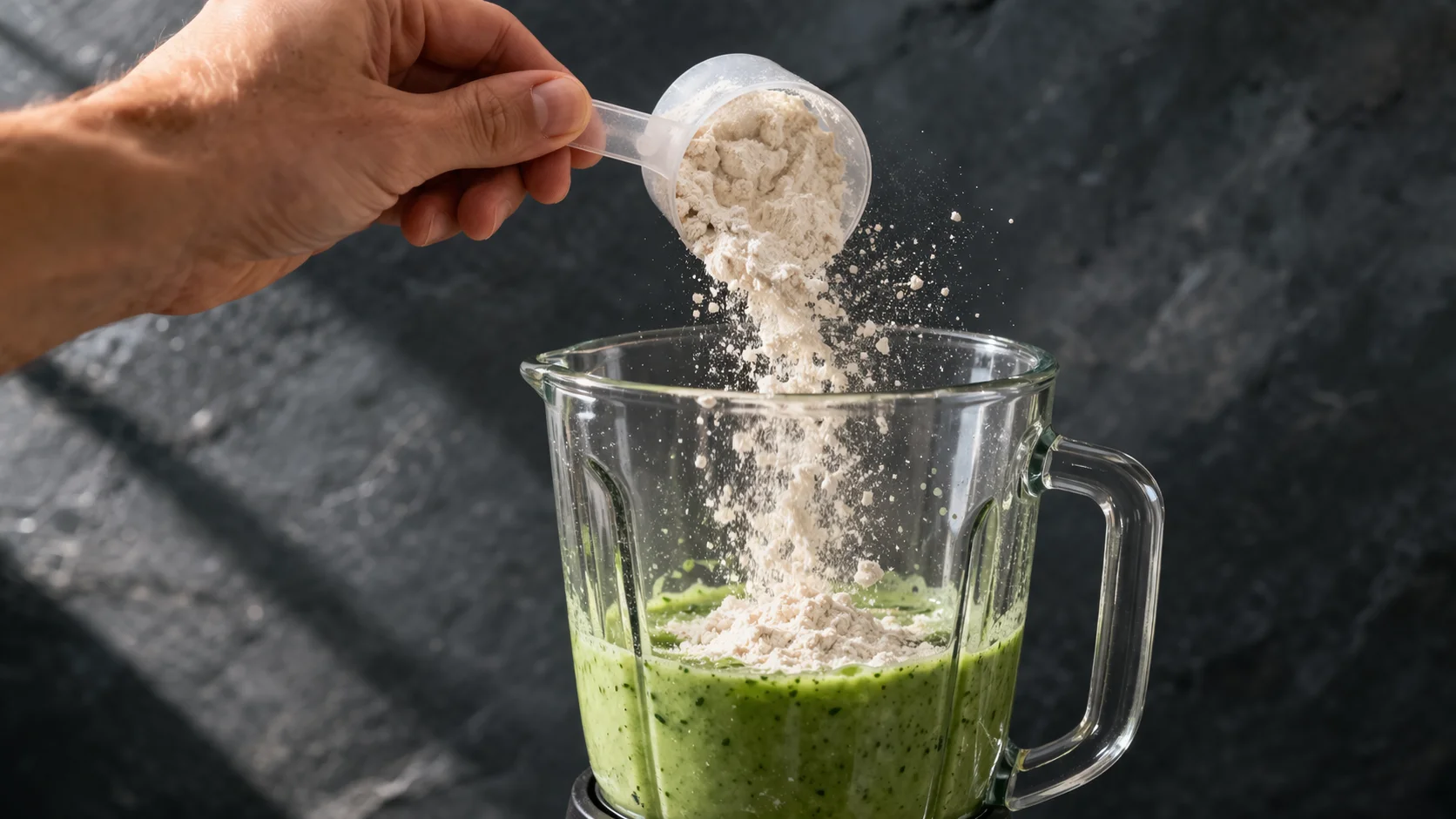 Main versant une mesure de protéine en poudre dans le blender contenant la base verte du smoothie