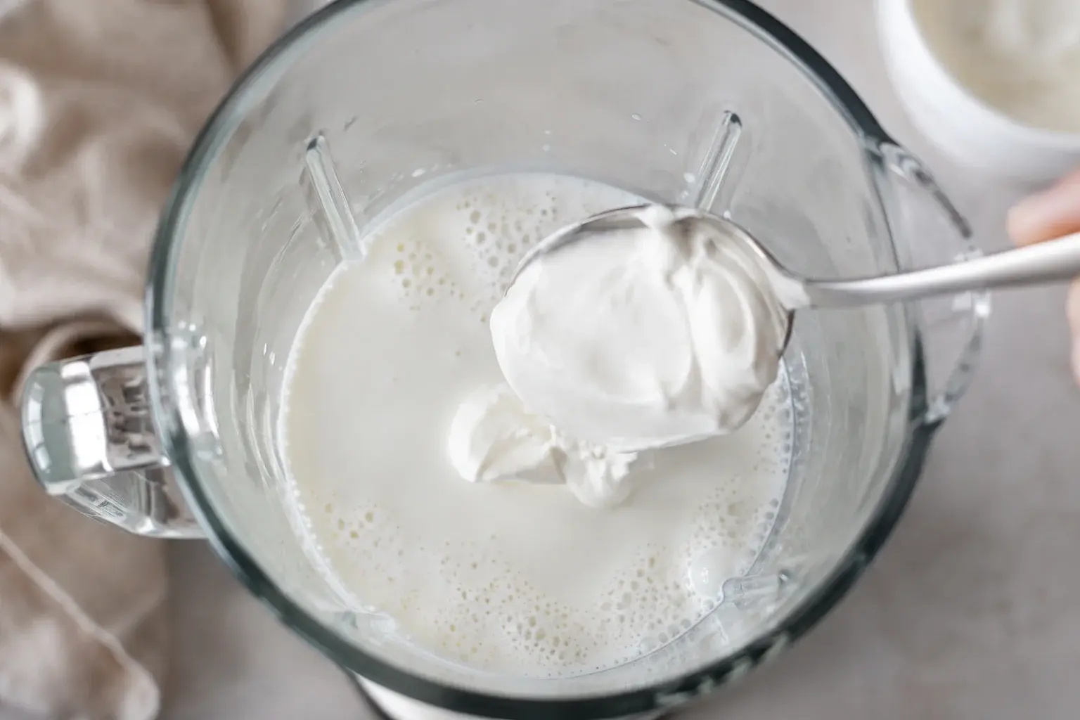 Yaourt grec blanc crémeux ajouté à la cuillère dans le bol transparent d'un blender contenant du lait, vue plongeante