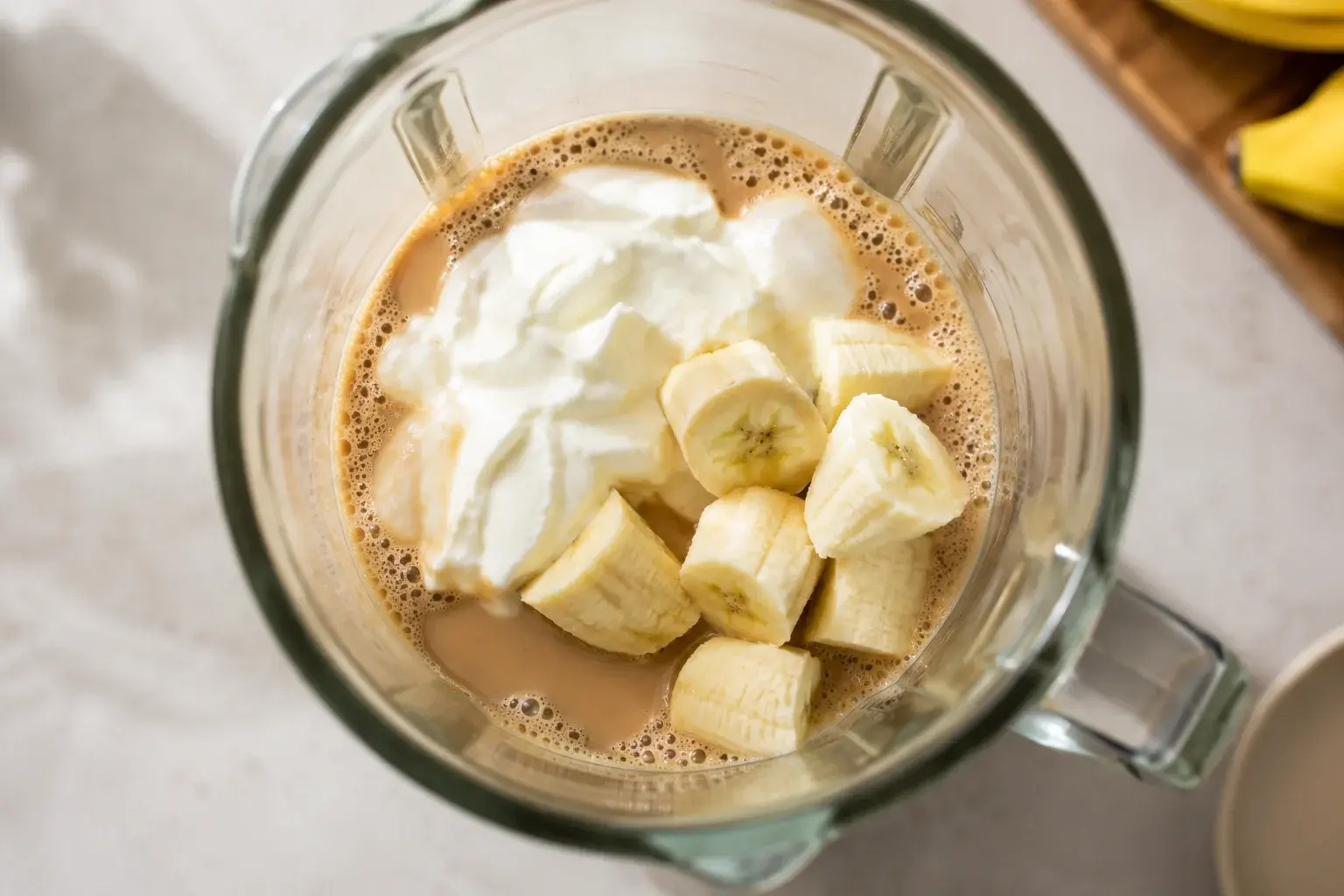 Yaourt grec blanc et tronçons de banane ajoutés dans un blender contenant du café brun et du lait, vue plongeante sur le bol
