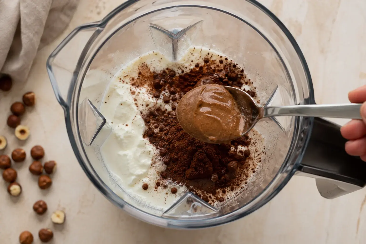 Cacao en poudre brun foncé et purée de noisette ajoutés dans un blender contenant du lait et du yaourt grec blanc, vue plongeante