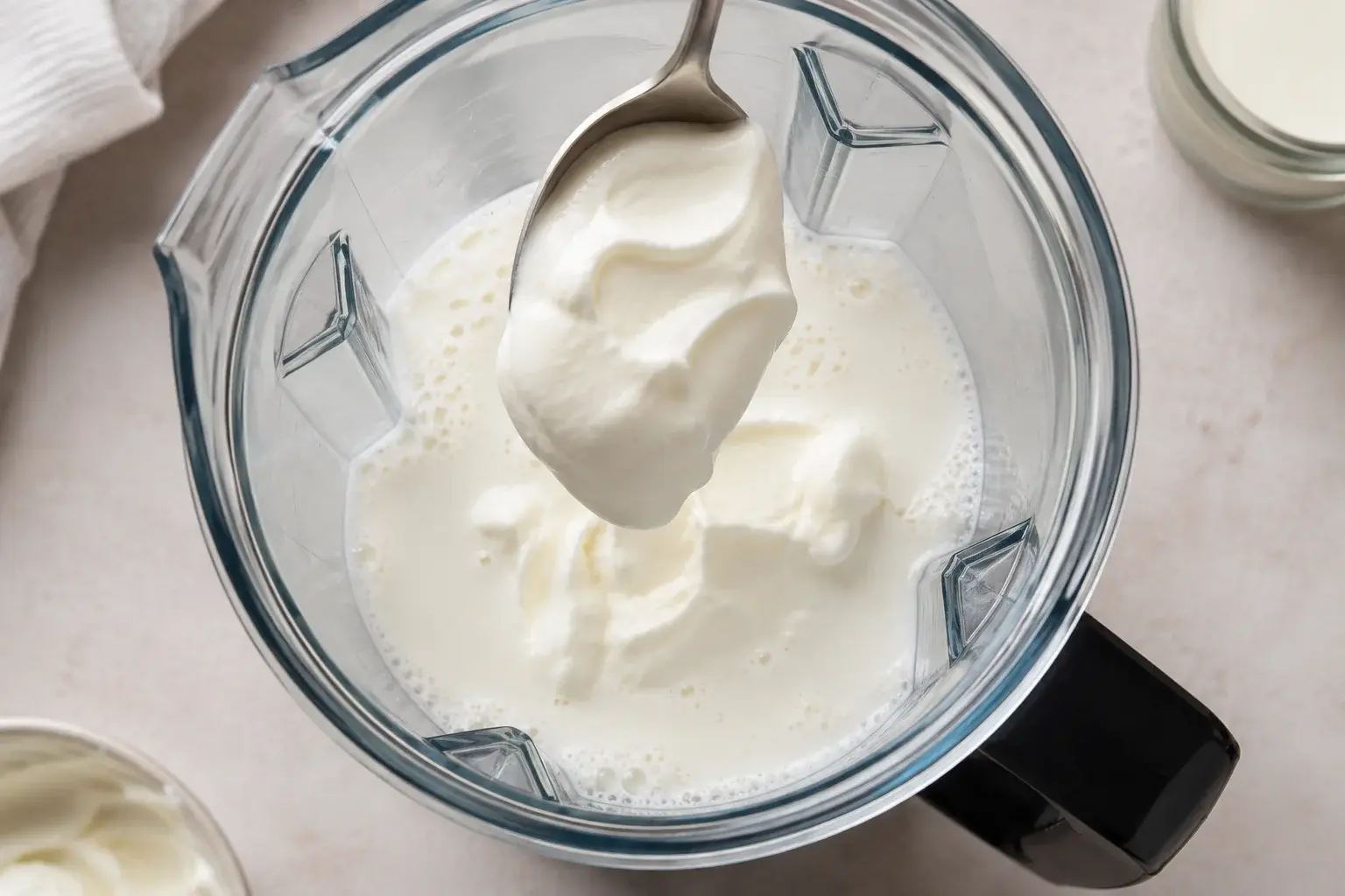 Yaourt grec blanc crémeux versé à la cuillère dans un blender contenant du lait blanc, vue plongeante sur le bol transparent