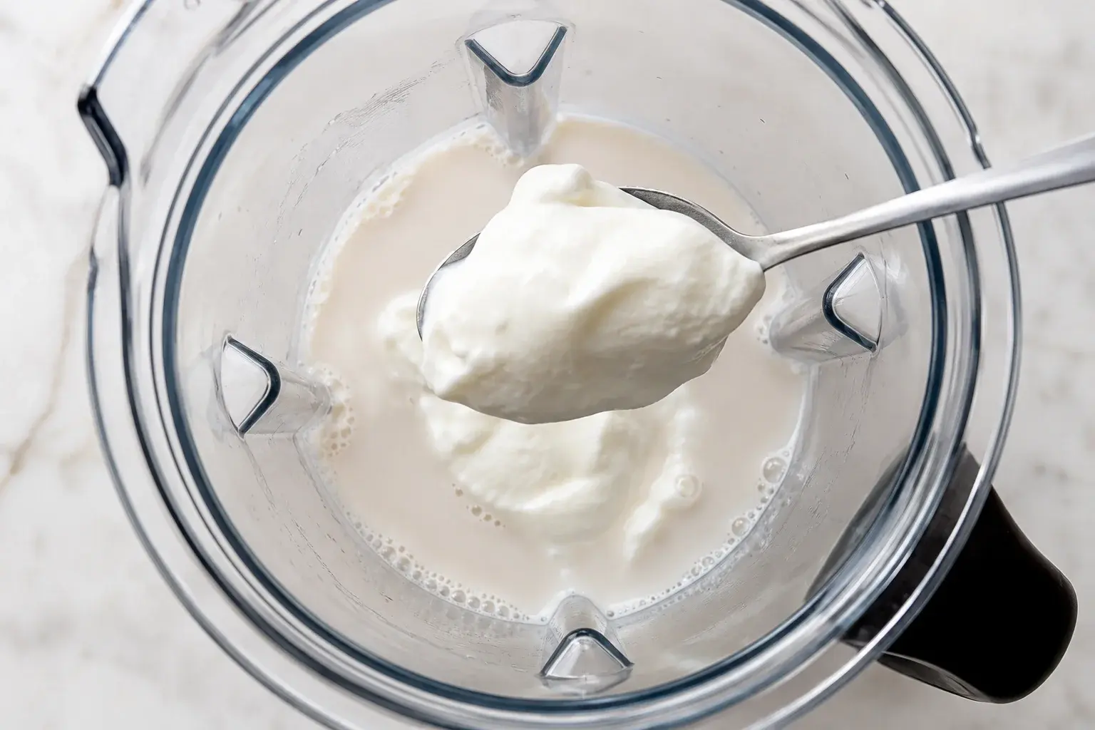 Yaourt grec blanc crémeux ajouté à la cuillère dans un blender contenant du lait d'amande blanc, vue plongeante sur le bol transparent