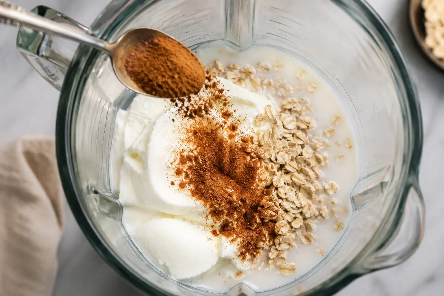Cannelle moulue brune saupoudrée dans le bol d'un blender contenant yaourt grec blanc, lait et flocons d'avoine, vue plongeante