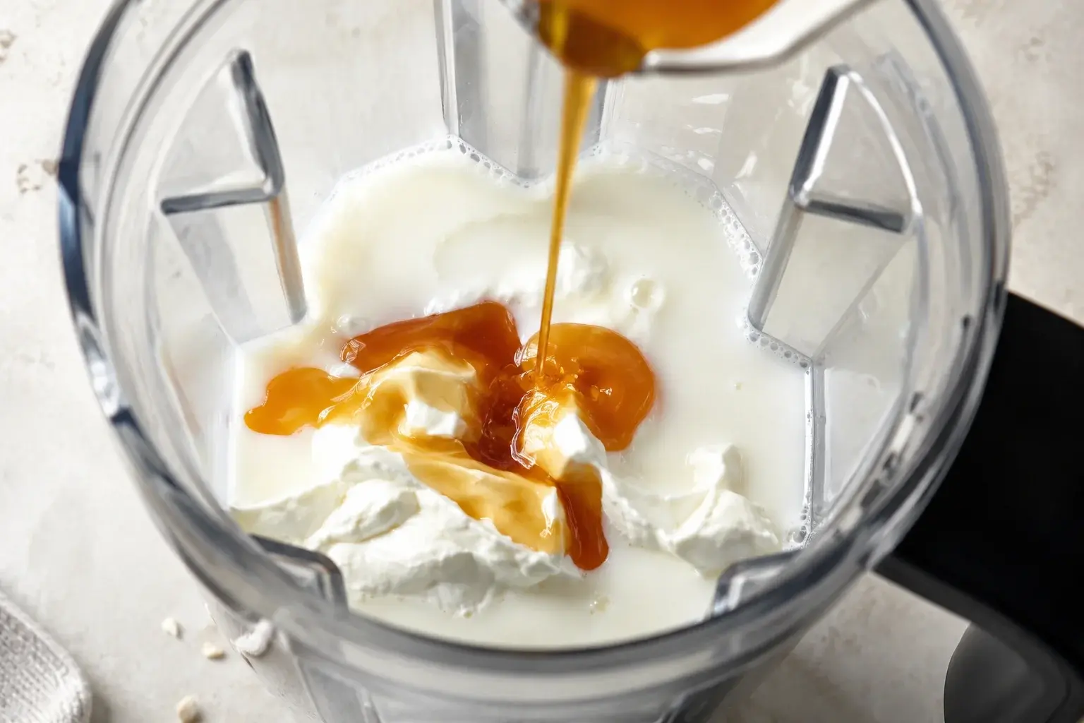 Sirop de caramel doré coulant dans le bol transparent d'un blender contenant yaourt grec blanc et lait, vue plongeante