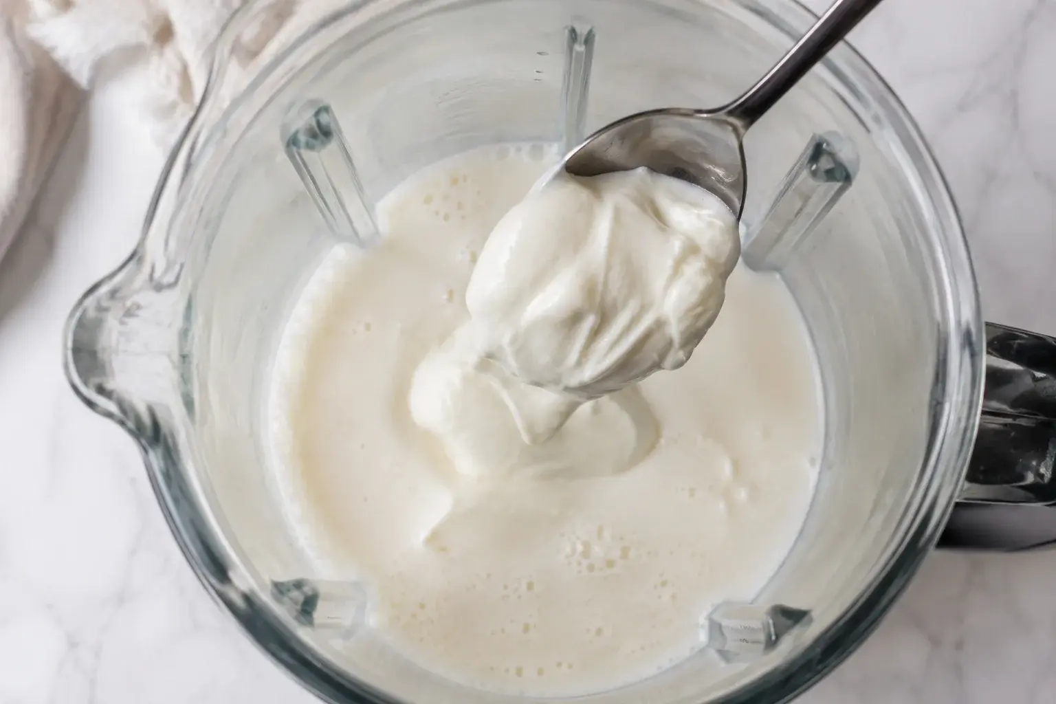 Yaourt grec blanc crémeux versé dans un blender contenant déjà du lait blanc, vue plongeante sur le bol transparent
