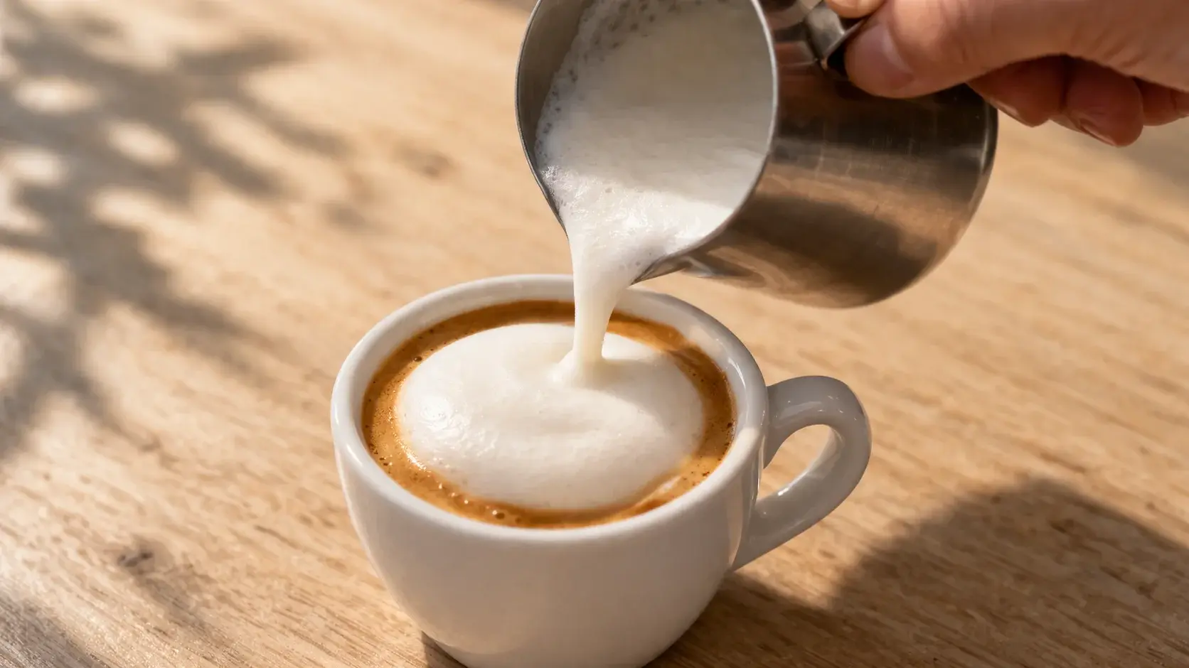 Main versant délicatement le lait protéiné chaud sur un shot d'espresso dans une tasse blanche, mousse crémeuse débordant sur le dessus, fond de bois clair