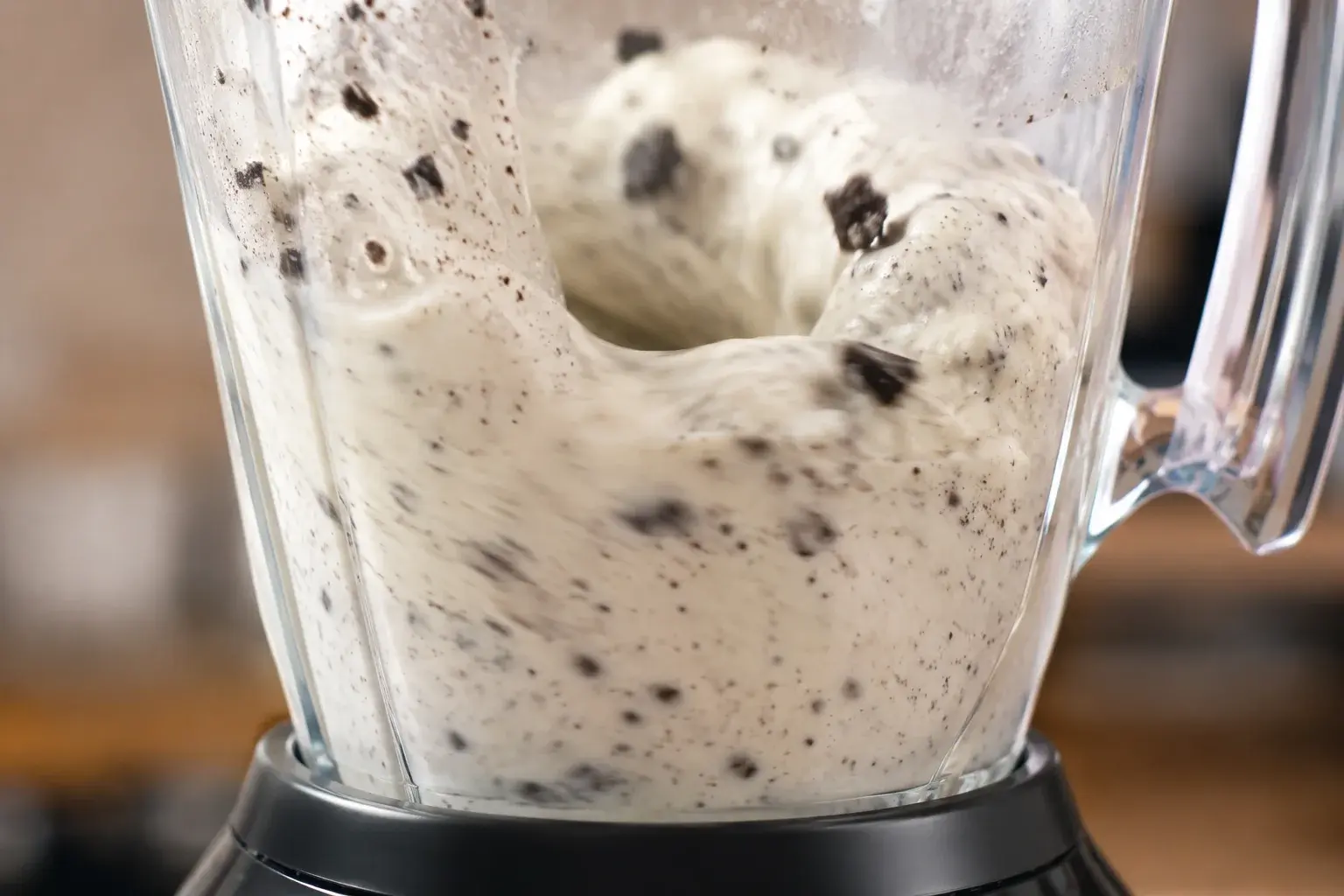 Blender en marche avec milkshake vanille blanc moucheté d'éclats de cookies noirs en train de tourbillonner derrière le bol transparent