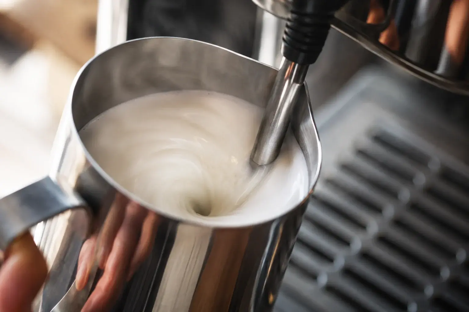 Buse vapeur en train de mousser du lait blanc dans un pichet en inox, tourbillon visible et microfoam soyeuse se formant en surface