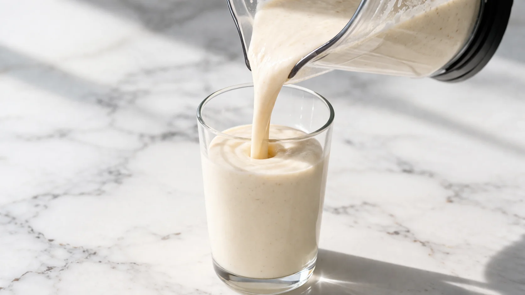 Verre de smoothie poire-vanille protéiné en cours de remplissage, liquide crémeux versé depuis le blender sur fond de marbre blanc