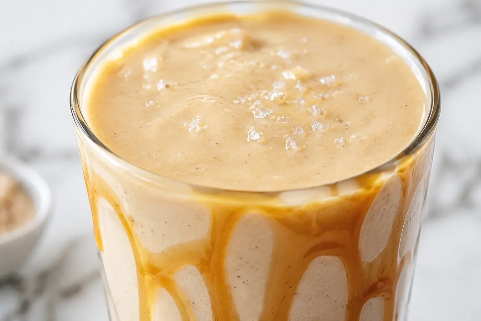 Gros plan sur un smoothie vanille-caramel beige onctueux dans un verre transparent, sirop de caramel coulant sur les bords et grains de fleur de sel