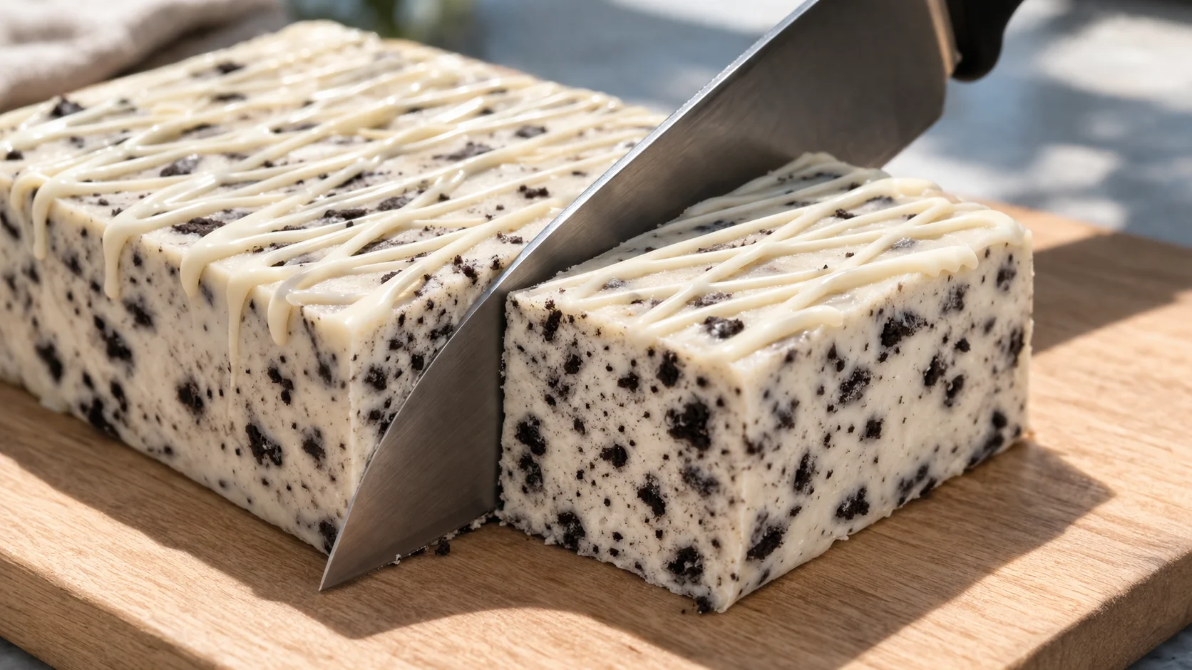 Grand couteau découpant le bloc de barres cookies and cream sur une planche en bois clair, coupe nette révélant la masse blanche aux éclats de biscuits noirs et le filet de chocolat blanc en surface