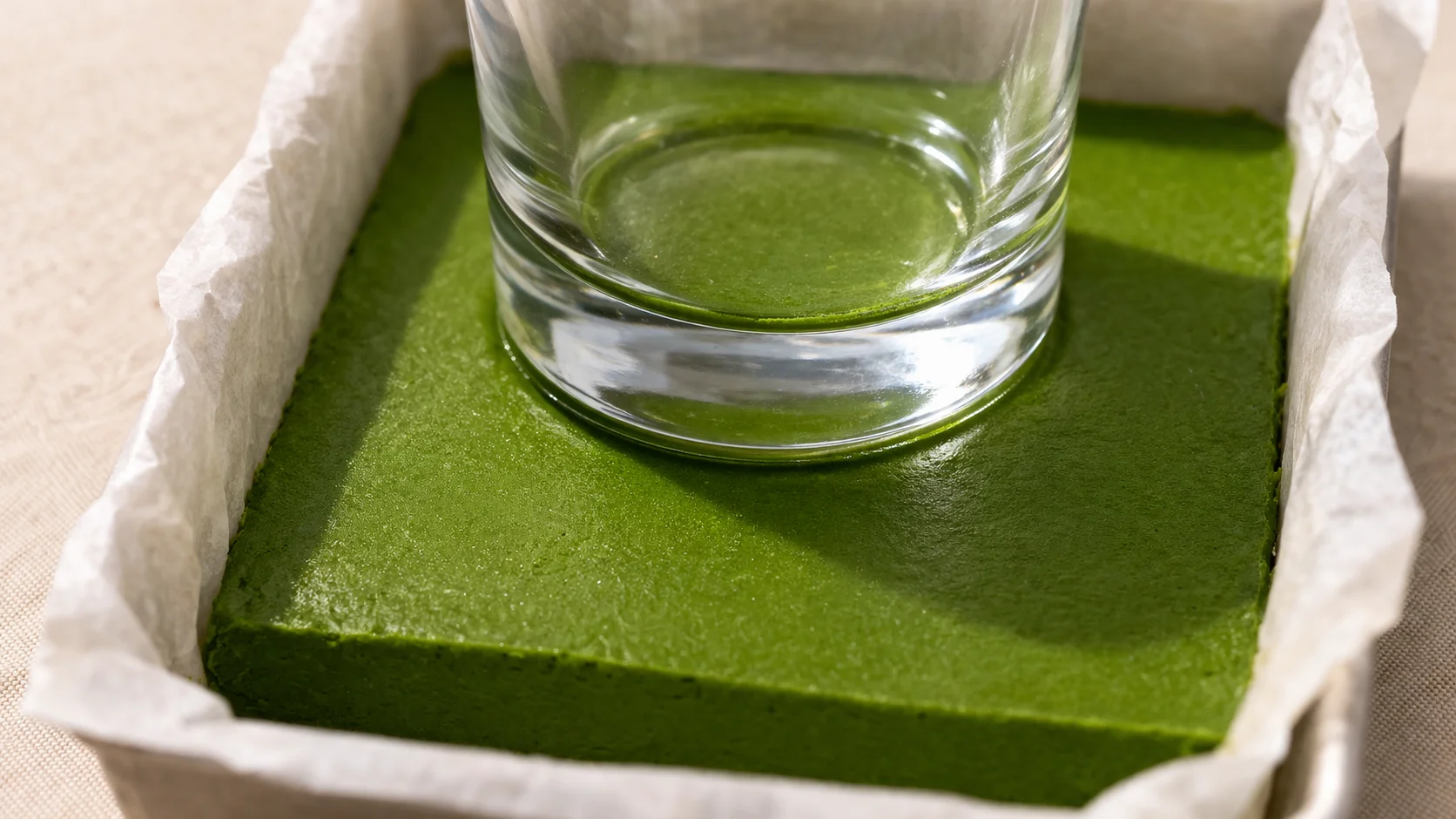 Fond de verre comprimant fermement la masse verte matcha dans un plat rectangulaire tapissé de papier cuisson, couleur vert intense et surface lisse bien visible, fond lin beige