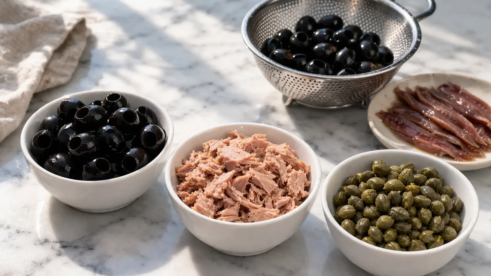 Olives noires dénoyautées, thon égoutté et câpres rincées disposés dans des petits bols sur un plan de travail en marbre blanc, passoire et filets d'anchois à côté