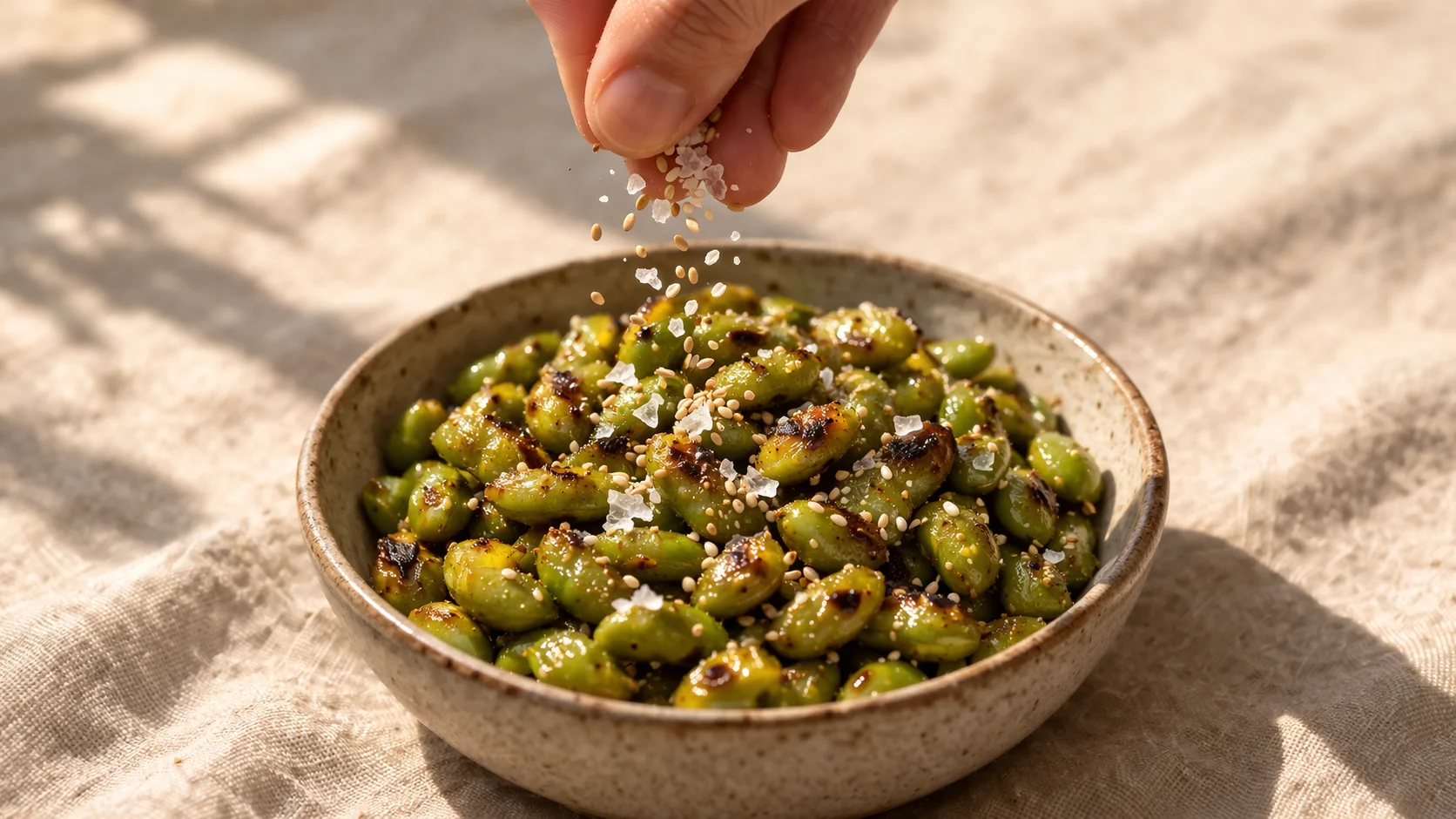 Bol en céramique blanche rempli d'edamame rôti épicé et doré, graines de sésame et fleur de sel saupoudrées sur le dessus, fond lin beige, lumière naturelle