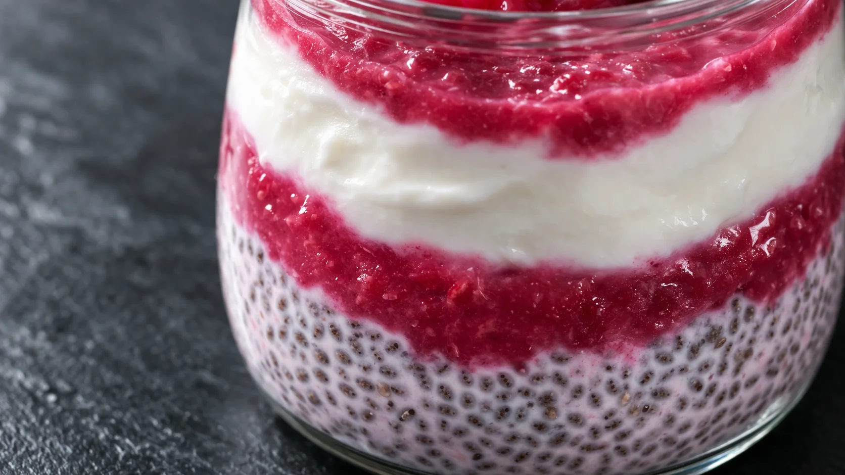 Gros plan sur la texture crémeuse d'un chia pudding framboise en bocal, couches de yaourt grec et coulis rose bien distinctes, graines de chia apparentes