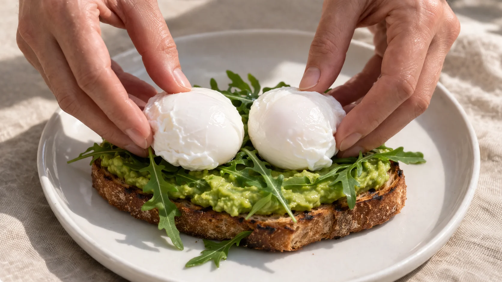 Deux œufs pochés en cours de dépôt sur une purée d'avocat verte étalée sur pain grillé, roquette visible en dessous, assiette en céramique blanche sur fond lin beige