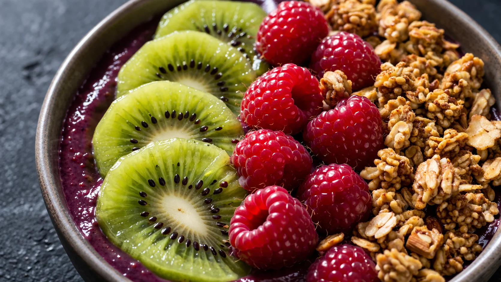 Gros plan sur les toppings colorés d'un bowl açaï, tranches de kiwi, framboises fraîches et granola croustillant sur base violette intense, fond ardoise
