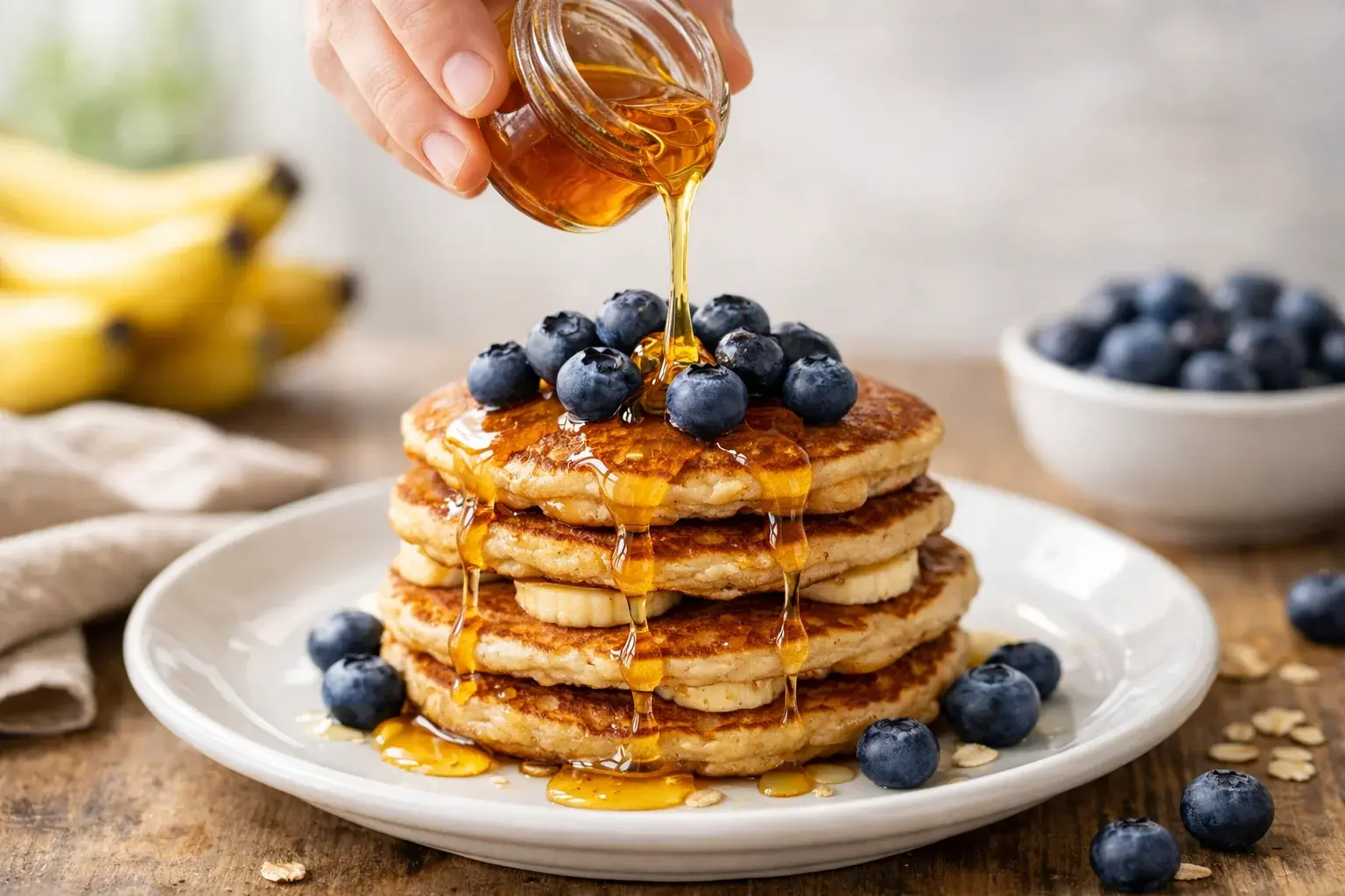 Main versant un filet de miel doré sur une pile de pancakes banane-avoine garnis de myrtilles fraîches, assiette blanche