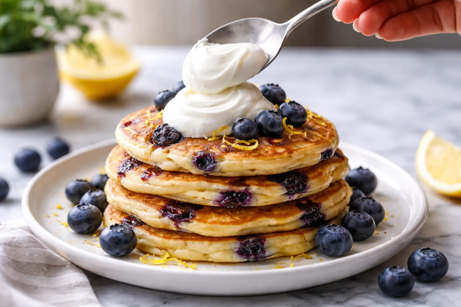 Main déposant une quenelle de yaourt grec sur une pile de pancakes myrtilles-citron, myrtilles fraîches et zestes de citron autour