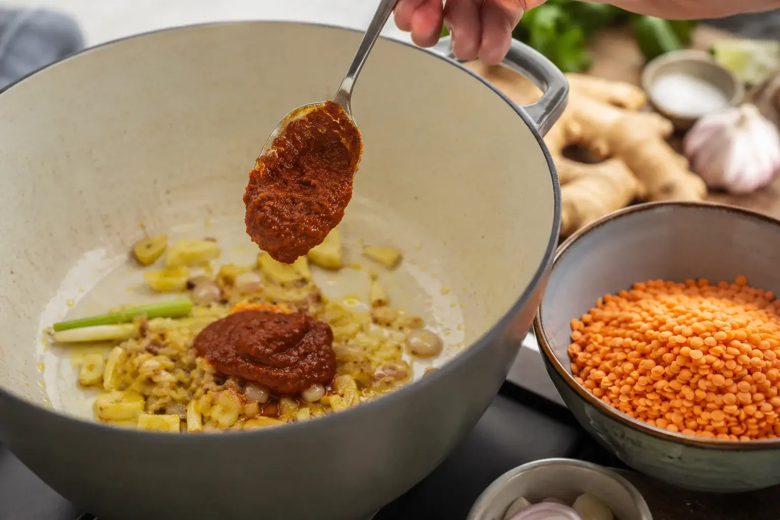 Pâte de curry rouge et épices en poudre versées sur les aromates en train de cuire, lentilles corail orange à côté