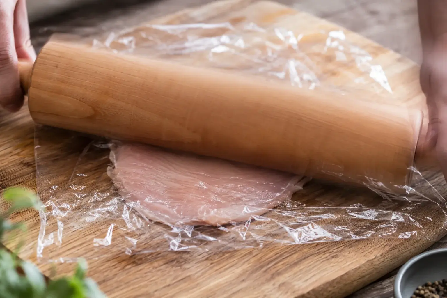 Rouleau à pâtisserie qui aplatit un blanc de poulet entre deux feuilles de film transparent sur une planche en bois