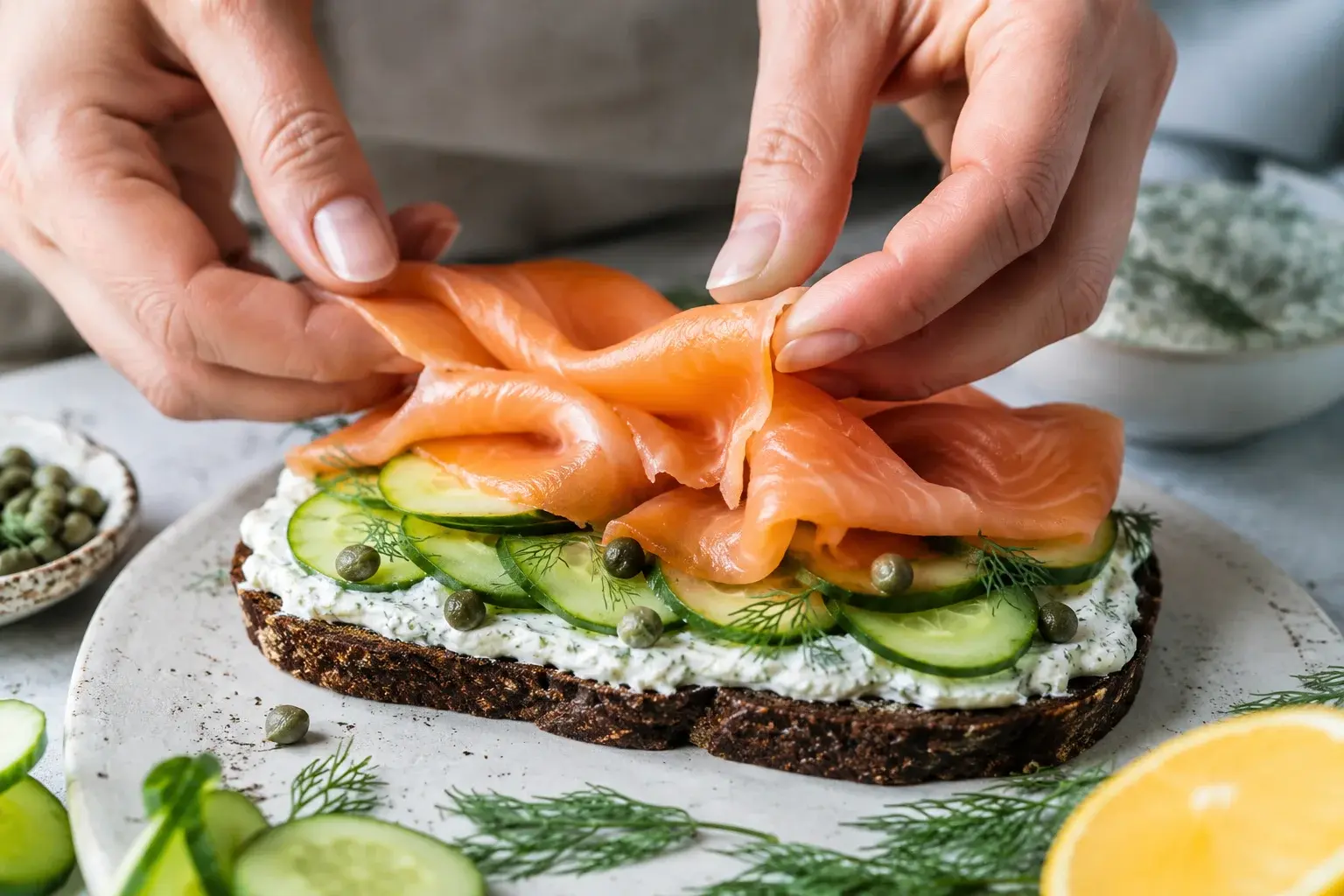 Mains qui déposent des tranches de truite fumée rose sur une tartine généreusement tartinée de fromage blanc, concombre et câpres visibles