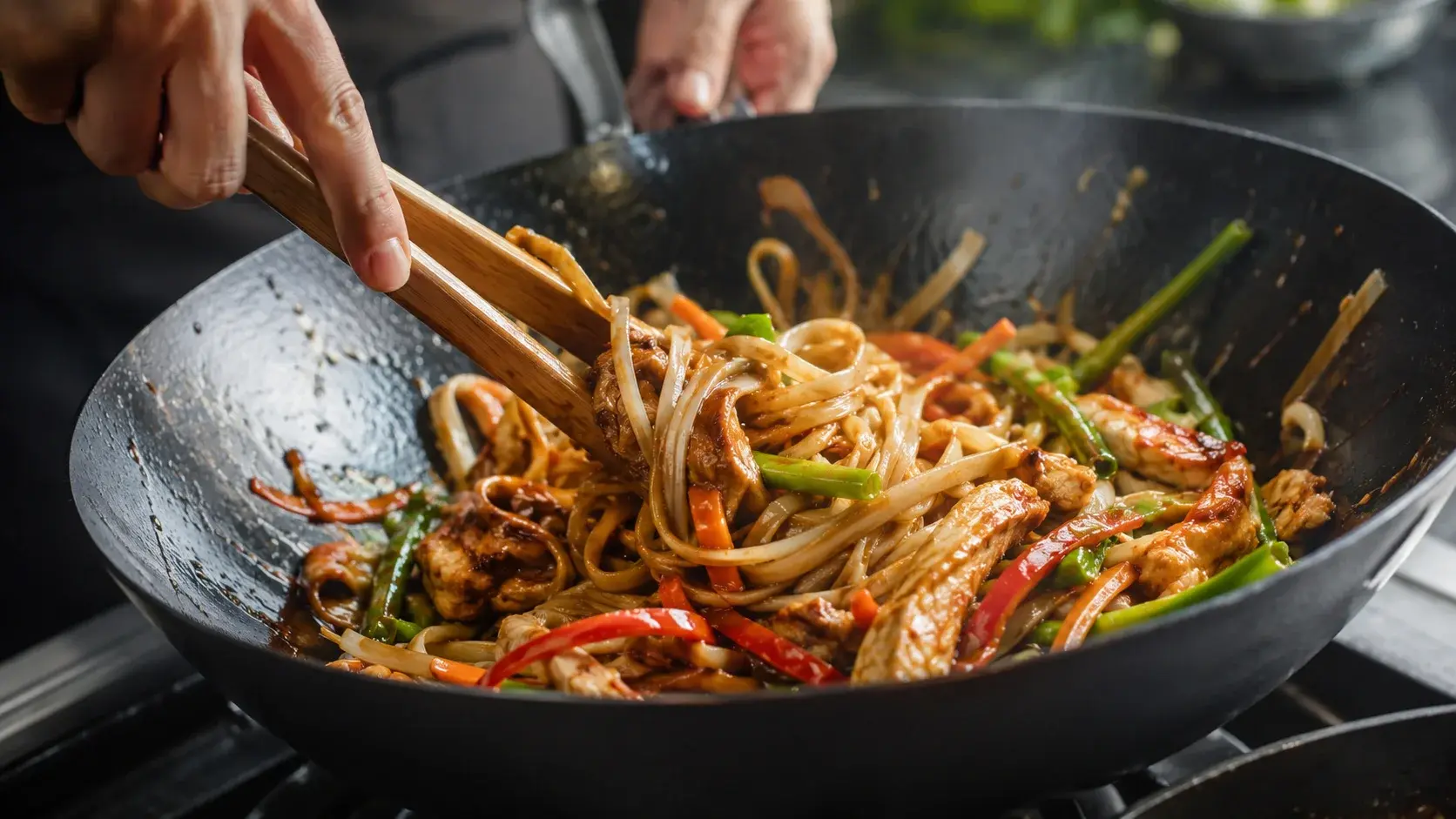 Mains tenant une pince qui mélange nouilles de riz, poulet et légumes colorés dans un wok avec sauce soja brillante