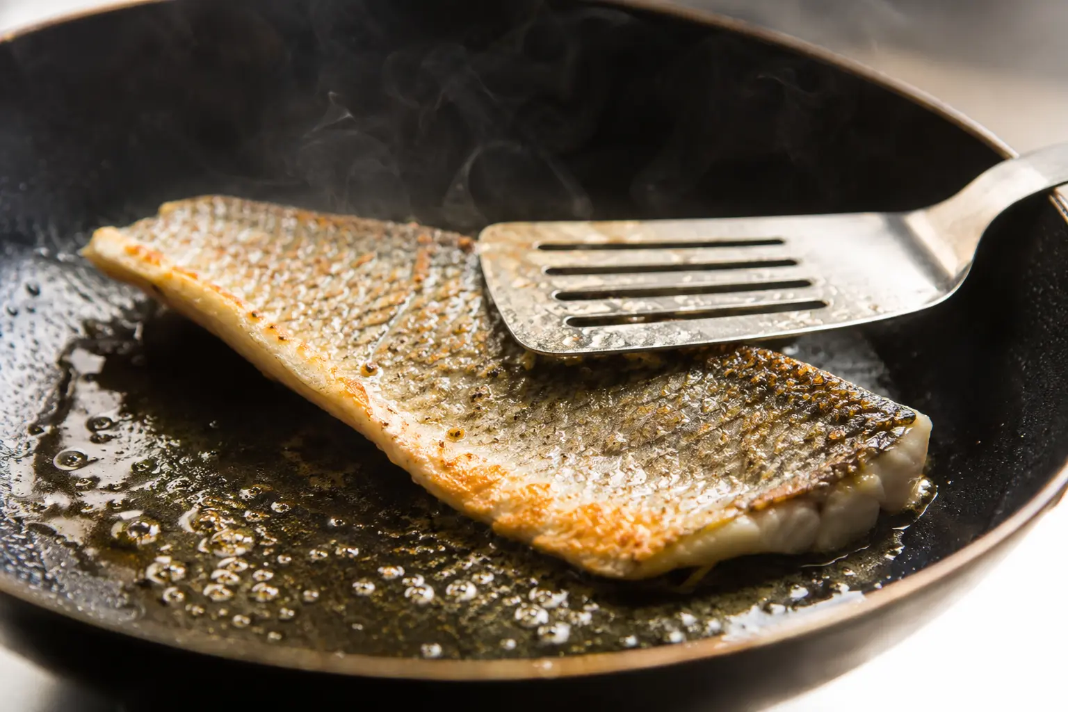 Filet de bar en train de griller côté peau dans une poêle chaude avec de l'huile d'olive fumante
