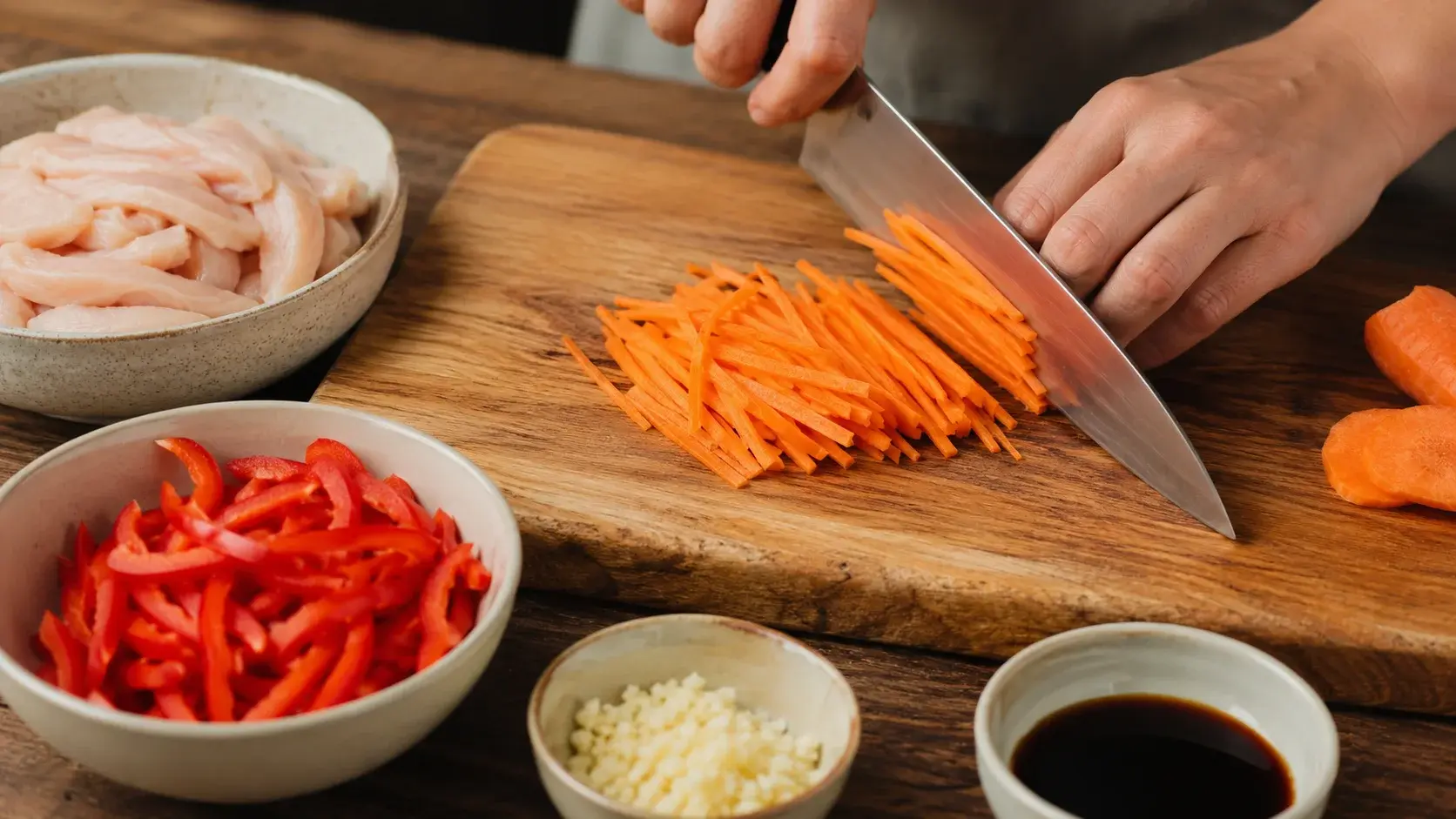 Plan de travail avec lanières de poulet, légumes colorés taillés en julienne et petit bol de sauce soja sur planche en bois