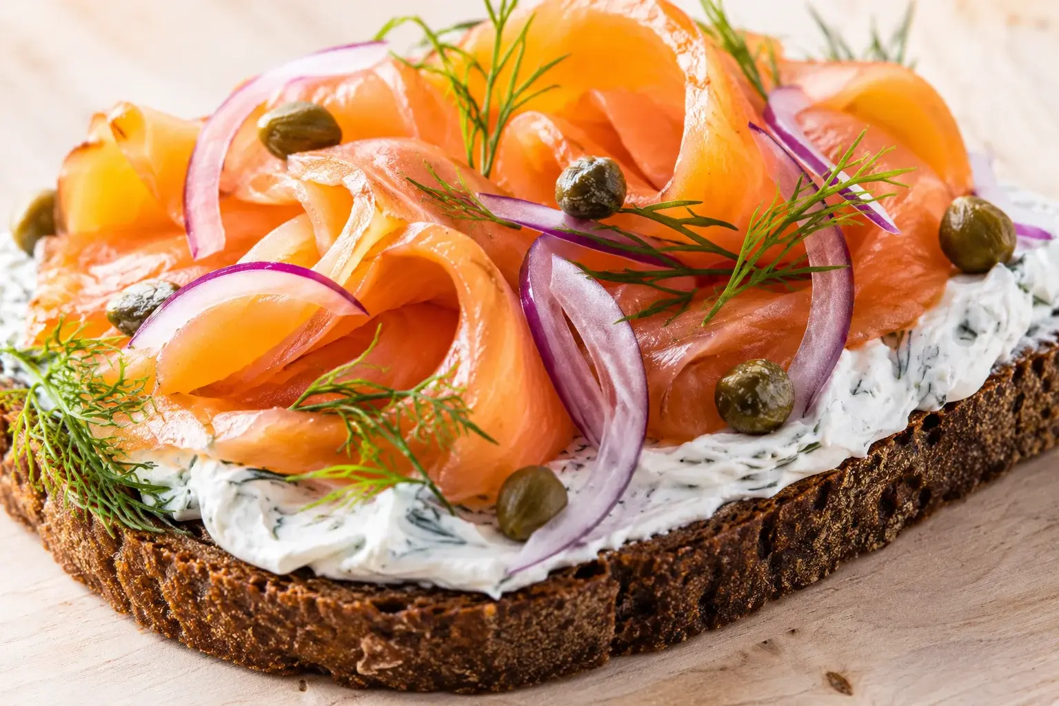 Gros plan sur une tartine de pain de seigle généreusement tartinée de fromage blanc à l'aneth, surmontée de truite fumée et câpres